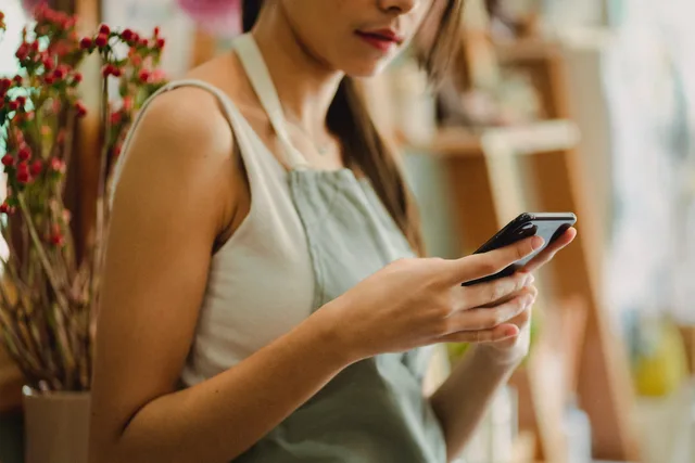 Crop anonymous florist in apron browsing mobile phone while working in cozy floral shop. Sociale medier for SMV'er: vælg kanaler og lav en plan